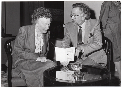 B&W photo of a man and a woman seated at a small round coffee table. The man has his face turned toward the woman while speaking to her. She is looking down at an event program, which he his holding in one hand. The program is titled, "Food and Fashions for the Field". In the background, a third person (partially obscured) is walking across the room behind them.