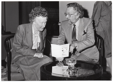 B&W photo of a man and a woman seated at a small round coffee table. The man has his face turned toward the woman while speaking to her. She is looking down at an event program, which he his holding in one hand. The program is titled, "Food and Fashions for the Field". In the background, a third person (partially obscured) is walking across the room behind them.