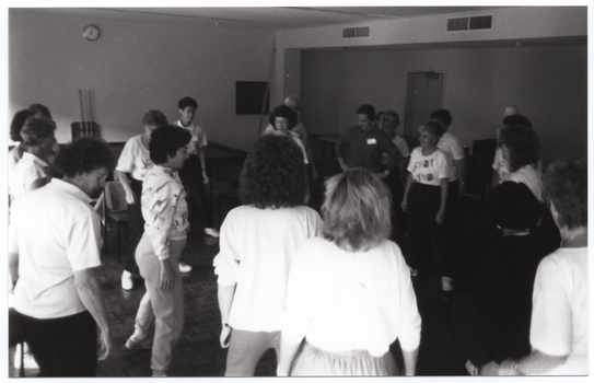 B&W photo of a group of women and one man (partially obscured) standing around a room in two circles (one inner circle and one outer circle). In the background, there is a plastic skeleton on a vertical stand (partially obscured) behind some of the women. There is a clock on one of the walls.