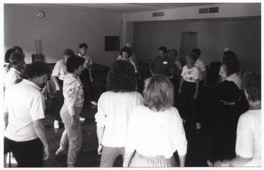 B&W photo of a group of women and one man (partially obscured) standing around a room in two circles (one inner circle and one outer circle). In the background, there is a plastic skeleton on a vertical stand (partially obscured) behind some of the women. There is a clock on one of the walls.
