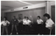 B&W photo of several women and one man (predominantly obscured) standing in two loosely defined semi-circles. They have their hands on their hips. Most of the women are wearing t-shirts with a "Joint Action Group" logo on the front. In the background, there is a plastic skeleton on a vertical stand (partially obscured). There are several bi-fold door panels folded against the wall in an open position.