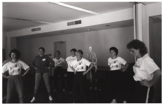 B&W photo of several women and one man (predominantly obscured) standing in two loosely defined semi-circles. They have their hands on their hips. Most of the women are wearing t-shirts with a "Joint Action Group" logo on the front. In the background, there is a plastic skeleton on a vertical stand (partially obscured). There are several bi-fold door panels folded against the wall in an open position.