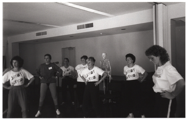 B&W photo of several women and one man (predominantly obscured) standing in two loosely defined semi-circles. They have their hands on their hips. Most of the women are wearing t-shirts with a "Joint Action Group" logo on the front. In the background, there is a plastic skeleton on a vertical stand (partially obscured). There are several bi-fold door panels folded against the wall in an open position.