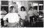 B&W photo of several women standing beside some large windows. Each person is standing on one leg, with one knee up while holding onto the back of a chair with one hand. Between the windows and the women, there is a tall metal-framed object, possibly some metal shelves or a food service trolley.