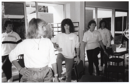 B&W photo of several women standing beside some large windows. Each person is standing on one leg, with one knee up while holding onto the back of a chair with one hand. Between the windows and the women, there is a tall metal-framed object, possibly some metal shelves or a food service trolley.