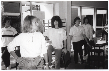 B&W photo of several women standing beside some large windows. Each person is standing on one leg, with one knee up while holding onto the back of a chair with one hand. Between the windows and the women, there is a tall metal-framed object, possibly some metal shelves or a food service trolley.