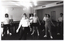 B&W photo of several women and one man standing in a large room. They have their arms outstretched by their sides, towards the floor. Behind them is a large billiard table, with some billiard balls on it. Against the wall, behind the billiard table, there are three pool cues on a stand and a wall clock on the wall above. Against another wall, there are two large tables on either side of a closed door. There is a chair in front of the door.