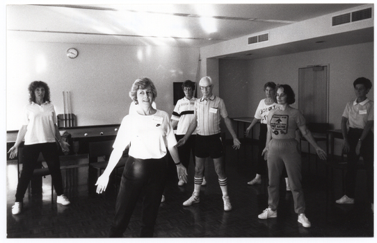 B&W photo of several women and one man standing in a large room. They have their arms outstretched by their sides, towards the floor. Behind them is a large billiard table, with some billiard balls on it. Against the wall, behind the billiard table, there are three pool cues on a stand and a wall clock on the wall above. Against another wall, there are two large tables on either side of a closed door. There is a chair in front of the door.