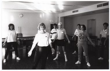B&W photo of several women and one man standing in a large room. They have their arms outstretched by their sides, towards the floor. Behind them is a large billiard table, with some billiard balls on it. Against the wall, behind the billiard table, there are three pool cues on a stand and a wall clock on the wall above. Against another wall, there are two large tables on either side of a closed door. There is a chair in front of the door.