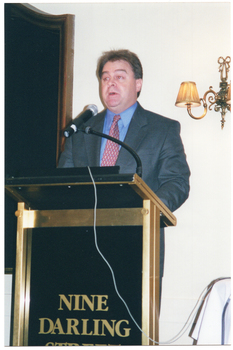 COL photo of a man, wearing a suit, shirt and tie. He is standing at a lectern and is speaking into a microphone, which is held by a short flexible arm attached to the lectern. On the front of the lectern, there is the text, "NINE DARLING". On the wall behind him, there is a large wooden-framed mirror on one side and a two-light sconce (wall lamp) on the other side.