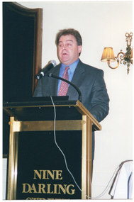 COL photo of a man, wearing a suit, shirt and tie. He is standing at a lectern and is speaking into a microphone, which is held by a short flexible arm attached to the lectern. On the front of the lectern, there is the text, "NINE DARLING". On the wall behind him, there is a large wooden-framed mirror on one side and a two-light sconce (wall lamp) on the other side.