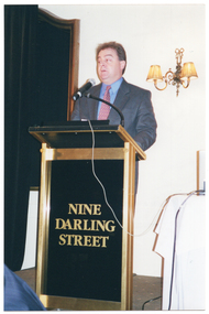 COL photo of a man, wearing a suit, shirt, tie, and spectacles. He is standing at a lectern and is speaking into a microphone, which is held by a short flexible arm attached to the lectern. On the front of the lectern, there is the text "NINE DARLING STREET". On the wall behind him, there is a large wooden-framed mirror on one side, partially obscured by a large black curtain. On the other side, there is a two-light sconce (wall lamp).