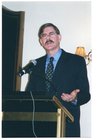 COL photo of a man, wearing a suit, shirt and tie. He is standing at a lectern. On the wall behind him, there is a large wooden-framed mirror on one side (partially obscured) and a sconce (wall lamp) on the other.