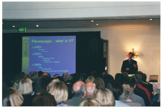 COL photo of a man, wearing a suit, shirt and tie. He is standing at a lectern. To his right, there is a large projector screen displaying a slide projection. The text heading on the slide says, "Fibromyalgia : what is it?", followed by dot points (illegible) under the sub-headings "1940's", "1970's", and "1980's". Above the screen, affixed to the ceiling, there is a small green "EXIT" sign. Behind the screen is a large black curtain. On the wall behind him, there is a large wooden-framed mirror on one side (partially obscured by the curtain) and a two-light sconce (wall lamp) on the other. There is a small table next to him covered with a white tablecloth, on top of which sits a laptop computer. In the foreground, there is a view of the audience from behind.