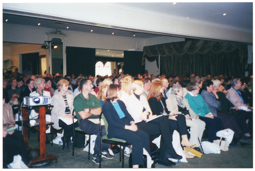 COL photo of a large group of people seated on several rows of chairs inside a large room. In the foreground, there is a slide projector on a wooden stand in the aisle between two sections of seating. There is a tall mirror in a decorative white wooden frame hanging on a narrow section of wall between two large openings, one of which has partially drawn black curtains. On another wall, there are large floor to ceiling olive green curtains decorated along the top with a draped French wave valance in a matching fabric.