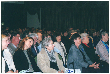 COL medium close-up photo of a large group of people, seated on several rows of chairs inside a large room. Several people have their heads leaning to one side. In the background, large floor to ceiling curtains are visible, decorated along the top with a draped valance.