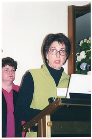 COL photo of a woman wearing eye glasses and a vest over a jumper. She is standing behind a lectern and is speaking into a microphone, which is held by a short flexible arm attached to the lectern. There is another woman standing behind her. On the wall behind her is a large wooden-framed mirror (partially obscurred), in front of which is a bouquet of flowers.