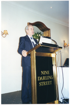 COL photo of a man, wearing a suit, shirt and tie. He is standing at a lectern and is speaking into a microphone, which is held by a short flexible arm attached to the lectern. On the wall behind him, there is a large wooden-framed mirror (partially obscured) with a two-light sconce (wall lamp) on the wall on either side of it. Beside him, there is a small table covered with a white tablecloth, on top of which sits a laptop computer.