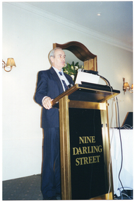 COL photo of a man, wearing a suit, shirt and tie. He is standing at a lectern and is speaking into a microphone, which is held by a short flexible arm attached to the lectern. On the wall behind him, there is a large wooden-framed mirror (partially obscured) with a two-light sconce (wall lamp) on the wall on either side of it. Beside him, there is a small table covered with a white tablecloth, on top of which sits a laptop computer.