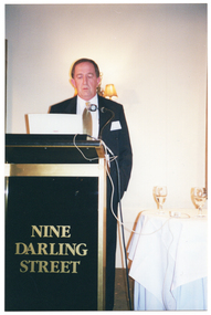 COL photo of a man, wearing a suit, shirt and tie. He is standing at a lectern and is speaking into a microphone, which is held by a short flexible arm attached to the lectern. On the front of the lectern, there is the text, "NINE DARLING STREET". On the wall behind him, there is a two-light sconce (wall lamp). Beside him, there is a small table covered with a white tablecloth, on which sit two glasses of white wine.