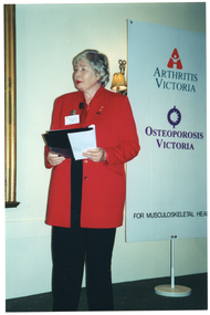COL photo of a woman, wearing a long jacket over trousers. She is speaking while holding an open folder. Behind her, there is a tall, freestanding vertical banner. It contains the text, "ARTHRITIS VICTORIA" and "OSTEOPOROSIS VICTORIA" and their respective logos, along with the text, "FOR MUSCULOSKELETAL HEALTH" (partially obscured). Beside the banner, affixed to the wall, is a two-light sconce (wall lamp), partially obscured).