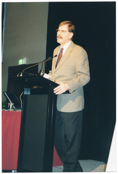 COL photo of a man standing at a lectern. He is speaking into a microphone attached to a flexible arm, which is held by a short flexible arm attached to the lectern. Beside him is a small table covered with a red tablecloth, on which sits a laptop computer and a glass of water. Behind him, there is a large floor to ceiling black curtain. In the background, to one side, there is a green "EXIT" sign above a doorway.