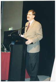 COL photo of a man standing at a lectern. He is speaking into a microphone attached to a flexible arm, which is held by a short flexible arm attached to the lectern. Beside him is a small table covered with a red tablecloth, on which sits a laptop computer and a glass of water. Behind him, there is a large floor to ceiling black curtain. In the background, to one side, there is a green "EXIT" sign above a doorway.