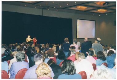 COL photo of two people standing on a large stage in the background, with a large seated audience in the foreground. Behind the people on the stage, there is a black floor to ceiling curtain which runs across the length of the stage. On the wall beside the stage is a large projector screen. Most of the people in the audience are seated, but there are a few people standing.