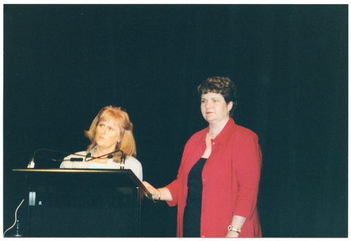 COL close-up photo of two women standing at a lectern. One woman is standing directly behind the lectern, which has two small microphones held by two flexible arms. She is wearing some light coloured clothing. The other woman is standing beside the lectern, with one hand placed on top. She is wearing a red jacket over some black clothing. Behind them is a large black floor to ceiling curtain.
