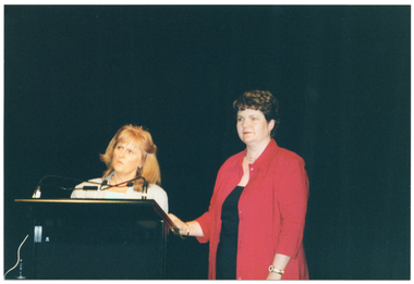 COL close-up photo of two women standing at a lectern. One woman is standing directly behind the lectern, which has two small microphones held by two flexible arms. She is wearing some light coloured clothing. The other woman is standing beside the lectern, with one hand placed on top. She is wearing a red jacket over some black clothing. Behind them is a large black floor to ceiling curtain.