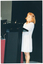 COL photo of a woman standing behind a lectern with two small microphones affixed to it. She is wearing a long light coloured striped jacket over a white dress. Beside her is a small table covered with a red tablecloth. Behind her, there is a large black floor to ceiling curtain.