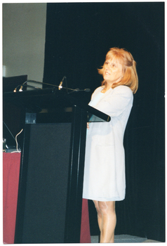COL photo of a woman standing behind a lectern with two small microphones affixed to it. She is wearing a long light coloured striped jacket over a white dress. Beside her is a small table covered with a red tablecloth. Behind her, there is a large black floor to ceiling curtain.