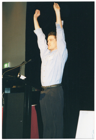 COL photo of a man standing on a stage, behind a lectern. He has both arms stetched up high above his head. Beside him is a small table covered with a red tablecloth. Behind him, there is a large black floor to ceiling curtain. In the background, there is a green "EXIT" sign above a doorway to the side of the stage.