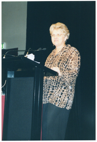 COL photo of a woman standing behind a lectern with two small microphones affixed to it. She is wearing a long brown patterned shirt over a black skirt. Beside her is a small table covered with a red tablecloth (partially obscured). Behind her, there is a large black floor to ceiling curtain. Off stage, there is a green "EXIT" sign above a doorway (partially obscured).