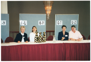 COL photo of four people sitting behind several long tables which have been joined together. The tables are covered with a white tablecloth with a long red valance around the sides. Behind them are three free-standing panels with white labels marked "E-H", "I-L", "M-P", "Q-T" and "U-Z". On the table, are three wrapped gifts. There is a large, modern, decorative chandelier hanging from the ceiling.