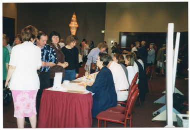 COL photo of a large group of people standing in a lobby. A few people are standing in front of some long tables, at which four people are seated. Behind the people seated are several freestanding display boards. In the background, there is a large modern decorative chandelier hanging from the ceiling.