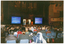 COL photo of a man standing at a lecturn, behind which are two free-standing vertical banners. On either side of him are two large screens displaying projected slides containing textual information. In the foreground, there is seated audience who have been Photographed from behind. In the background, there are several sections of wood panelling and several organ pipes separated into five groups.
