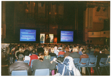 COL photo of a man standing at a lecturn, behind which are two free-standing vertical banners. On either side of him are two large screens displaying projected slides containing textual information. In the foreground, there is seated audience who have been Photographed from behind. In the background, there are several sections of wood panelling and several organ pipes separated into five groups.