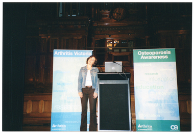 COL photo of a woman standing beside a lectern on a stage. She is tilting her head towards her left shoulder while holding her arms straight down by her sides. Behind her are two vertical free-standing banners, positioned on either side. One banner is titled 'Osteopororis Victoria' and the other is titled 'Osteoporosis Awareness'. In the background, there are several sections of wood panelling and a clock above an organ.