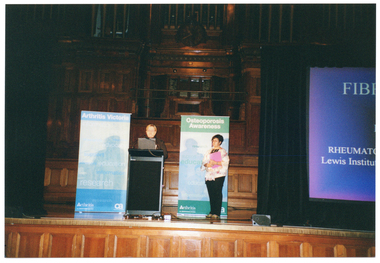COL photo of two women standing on a stage. One of the women is behind a lectern. Behind them, on either side of the lectern, are two free-standing vertical banners, titled 'Osteopororis Victoria' and 'Osteoporosis Awareness'. There is a large screen to one side (partially obscured), onto which a slide containing textual information is being projected. In the background, there are several sections of wood panelling and a clock above an organ.
