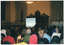 COL photo of a woman standing at a lectern, speaking into a microphone. Behind her, there is a small screen positioned to one side in front of a decorative section of wood pannelling on the wall. On either side, there are two large flags on the wall. In the foreground, there is a seated audience who have been Photographed from behind.