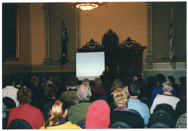 COL photo of a woman standing at a lectern, speaking into a microphone. Behind her, there is a small screen positioned to one side in front of a decorative section of wood pannelling on the wall. On either side, there are two large flags on the wall. In the foreground, there is a seated audience who have been Photographed from behind.