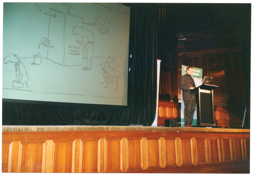 COL photo of a man speaking into a microphone at a lectern on a stage. On either side of him are two vertical free-standing banners (partially obscured). One of the banners contains the text "Osteoporosis Awareness" (partially obscured). On the stage, there is a large screen on a black floor to ceiling curtain, behind which there are some tall organ pipes. A slide of a Micheal Leunig cartoon is being projected onto the screen. In the cartoon, some text on a simple depiction of a large machine says, "XEROX THOUGHT SHREDDER".