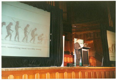 COL photo of a man speaking into a microphone at a lectern on a stage. On either side of him are two vertical free-standing banners (partially obscured). One of the banners contains the text "Osteoporosis Awareness" (partially obscured). Behind the man is a large screen on a black floor to ceiling curtain. The image being projected onto the screen contains several depictions of an upright neanderthal man holding various tools such as a knife or a spear. In the final depiction, the neanderthal man is sitting hunched over at a desk. Below the image is the text [partially obscured], ..."ere, something went terribly wrong".