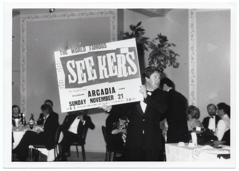 B&W photo of a man holding up an advertising billboard.  The text on the billboard is, "THE WORLD FAMOUS SEEKERS BALLROOM ARCADIA CORK SUNDAY NOVEMBER 21 9-2 7/8". There is some additonal text in smaller font which is illegible. Placed around various points on the billboard are the song titles "I'll Never Find Another You", "In A World Of Our Own", and "The Carnival Is Over". In the background, there are groups of people seated at round tables.