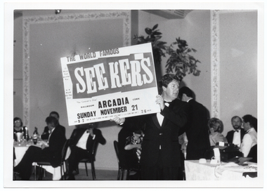 B&W photo of a man holding up an advertising billboard.  The text on the billboard is, "THE WORLD FAMOUS SEEKERS BALLROOM ARCADIA CORK SUNDAY NOVEMBER 21 9-2 7/8". There is some additonal text in smaller font which is illegible. Placed around various points on the billboard are the song titles "I'll Never Find Another You", "In A World Of Our Own", and "The Carnival Is Over". In the background, there are groups of people seated at round tables.