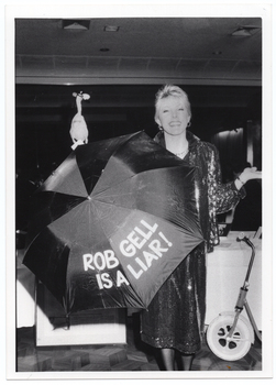 B&W photo of a woman holding up an open dark coloured umbrella. The words "ROB GELL IS A LIAR" have been written on the umbrella in large text. Behind her is a long table covered with a tablecloth. There is a scooter leaning against the table. Behind and above the umbrella, there is a plastic chicken.