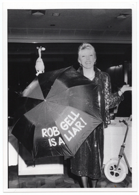 B&W photo of a woman holding up an open dark coloured umbrella. The words "ROB GELL IS A LIAR" have been written on the umbrella in large text. Behind her is a long table covered with a tablecloth. There is a scooter leaning against the table. Behind and above the umbrella, there is a plastic chicken.