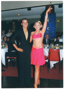 COL photo of a young couple, dressed in ballroom dancing costumes, posing for a photo at an event. Behind them, various people are either seated at round tables or standing. The tables are covered in white tablecloths, and there are platters of sandwiches, glasses, and cups and saucers on top. In the background, there is a blue and gold banner, with Arthritis Victoria and Osteoporosis Victoria text and logos, hanging from the ceiling.