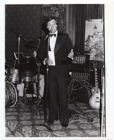 B&W photo of a man standing on stage at a microphone on a stand. He is holing a small piece of paper in one hand. Behind him, there is a drummer (partially obscured) sitting behind a drum kit, and an electric bass guitar leaning against some sound equipment (amplifiers).